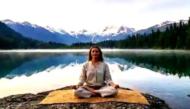एक महिला शांत झील के पास 'ध्यान' (Meditation) मुद्रा में बैठी है, जो मन को शांत करने की कला को दर्शाती है I (A woman in a peaceful environment sitting in a meditation posture by a calm lake, illustrating the 'Dhyan' (Meditation) keyword and the concept of calming a turbulent mind.)
